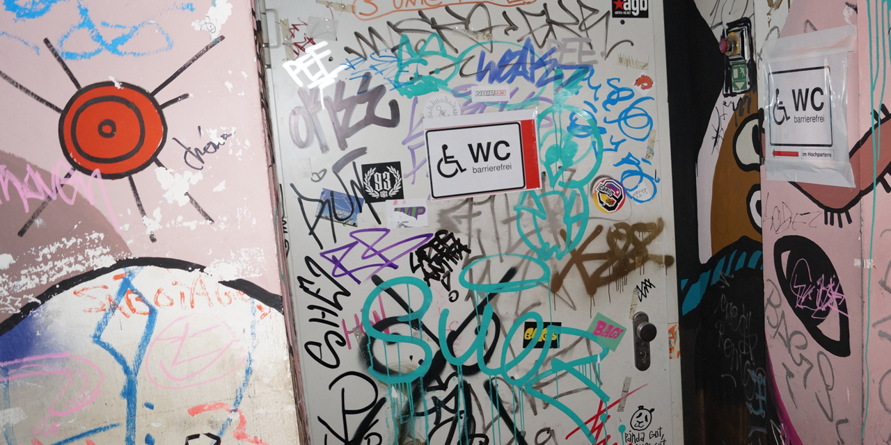 Das Bild zeigt die Tür der barrierfreien Toilette. Zwei Schilder mit Rollstuhl-Signet weisen darauf hin. Die Tür und die Wand sind mit Graffitti verziert.