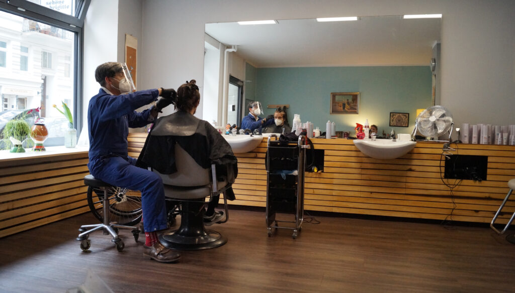 Auf dem Bild ist links ein Friseur zu sehen, der seinem Kunden die Haare schneidet. in der rechten Bildhälfte ist der Platz vor dem Waschbcken frei, hier könnte ein Elektrorollstuhl Platz finden.