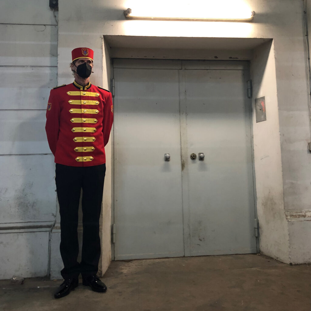 Ein Liftboy in rot-schwarzer Uniform mit goldenen Applikationen steht vor einer grauen Aufzugtür.