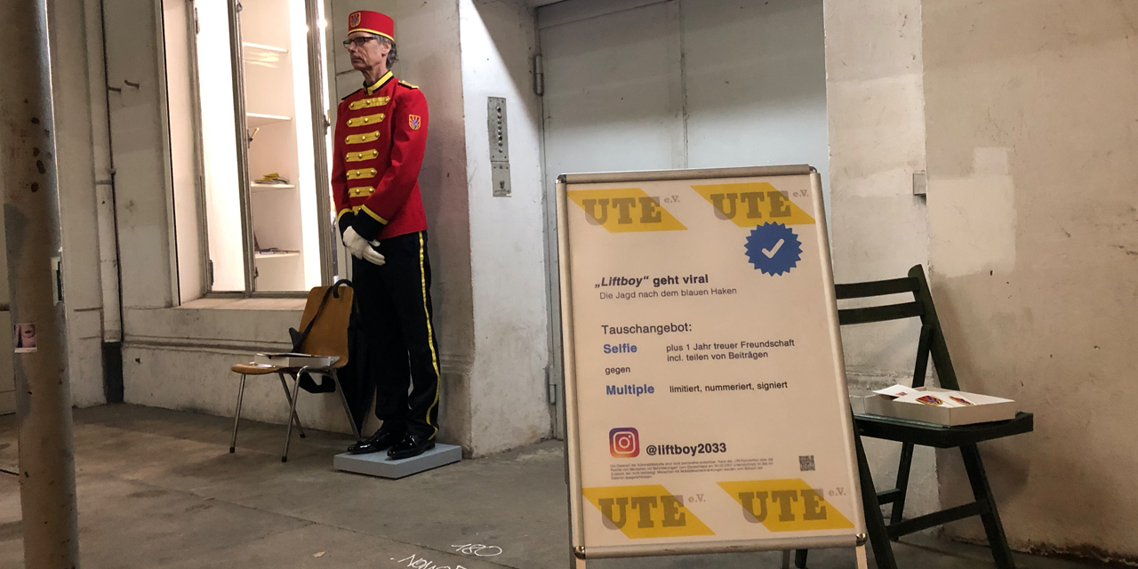 Ein Mann in Liftboy-Uniform steht vor einem Fahrstuhl, davor ein Plakat-Aufsteller.