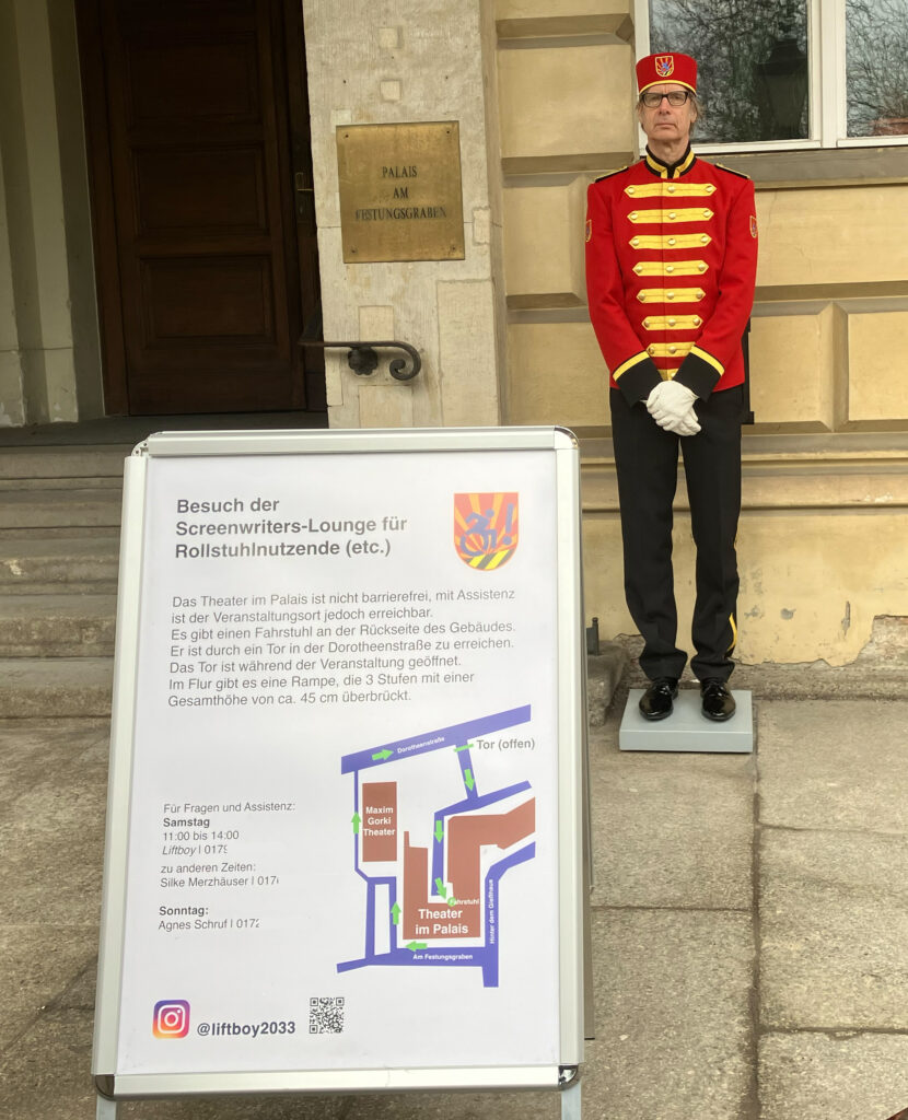 Ein Schild beschreibt die Zugangssituation für Rollstuhlnutzende, im Hintergrund ein Mann in Liftboy-Uniform